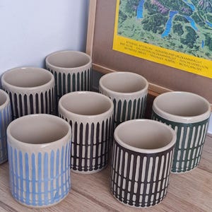 Op de afbeelding: Een verzameling keramische mokken met een beige binnen- en buitenkant. De buitenkant heeft verticale streepontwerpen in verschillende kleuren, waaronder blauw, groen en bruin. Op de achtergrond is een ingelijste kaart te zien.
