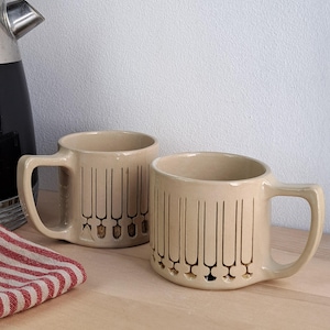 Peut inclure: Deux mugs en céramique beige avec un motif de lignes verticales et de petites formes en forme de pelle en bas. Les mugs ont de grandes anses et sont posés sur une surface en bois clair. Un tissu rayé rouge et blanc est visible à gauche.