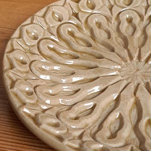 Può includere: Un piatto in ceramica beige con un motivo floreale in rilievo e ornato. La superficie del piatto ha una finitura lucida e il disegno è composto da forme organiche e vorticose. Il piatto è appoggiato su una superficie di legno.