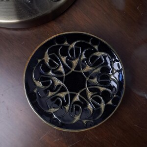 May include: A black and gold decorative coaster with an intricate, swirling design. The coaster is round and has a glossy finish. The design features a repeating pattern of curved lines and shapes. The coaster is likely made of ceramic or a similar material.