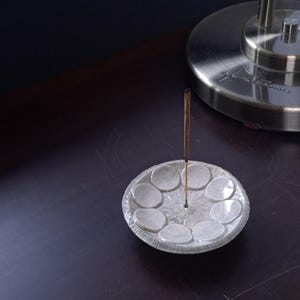 Peut inclure: Un porte-encens en verre transparent avec un design circulaire, comportant un trou central pour le bâton d'encens et des renfoncements circulaires environnants. Un bâton d'encens allumé est au centre. Un pied de lampe argenté est en arrière-plan.