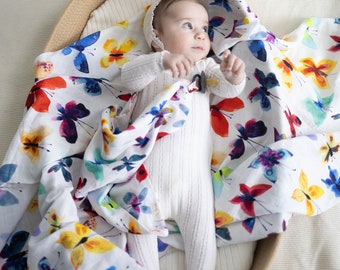 May include: A baby wearing a white onesie lies in a woven basket, swaddled in a white muslin swaddle blanket with a colorful watercolor butterfly print.