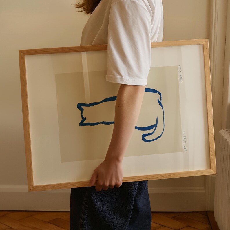 May include: A framed art print featuring a minimalist blue cat illustration on a cream background. The artwork is encased in a light wood frame. Text on the print reads "CAT. LINE 01" and "Catch some".