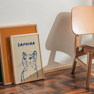 K&ouml;nnte beinhalten: Drei gerahmte Kunstdrucke lehnen an einer Wand. Der zentrale Druck zeigt eine blaue Katzenillustration mit dem Namen "SAPHIRA" dar&uuml;ber. Die Rahmen sind aus hellem Holz, mit einem gr&uuml;nen und orangefarbenen Druck, der hinter dem Katzendruck sichtbar ist. Rechts ist ein Holzstuhl.