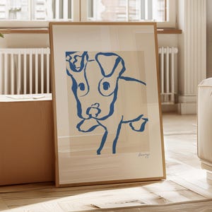 May include: A framed art print with a minimalist blue line drawing of a dog. The artwork is set against a cream background and framed in light wood. The print is leaning against a cardboard box in a well-lit room.