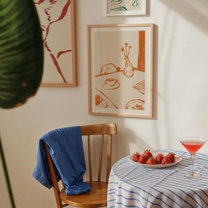 May include: A framed print with an orange and white illustration of a table setting, a blue jacket draped over a wooden chair, and a table with a plate of strawberries and a cocktail. The print is in a light wood frame.