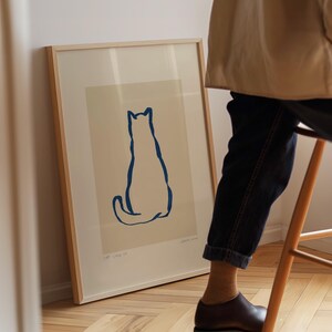 May include: A framed art print featuring a minimalist blue outline of a cat on a cream background. The artwork is displayed in a light wood frame, leaning against a wall. The print includes the text "CAT. Still life".
