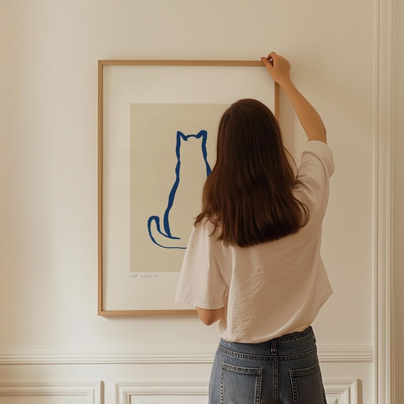May include: A framed art print featuring a minimalist blue cat silhouette on an off-white background. The artwork is enclosed in a light brown wooden frame. The print is being hung on a white wall.