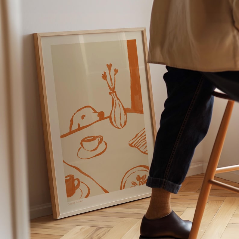 May include: A framed art print featuring an orange and cream-colored illustration. The artwork depicts a table setting with a dog, coffee cups, a vase with flowers, and a plate. The print is in a light wood frame.