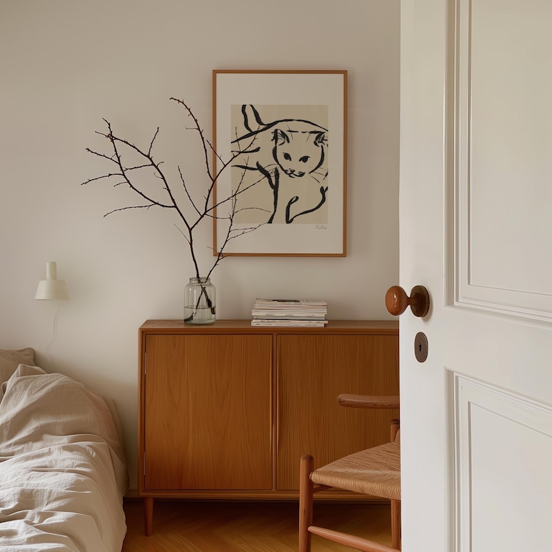 May include: A minimalist room scene with a framed cat print, a wooden cabinet, and a glass vase holding branches. The artwork is in black and white. A bed and a chair are partially visible, adding to the room's aesthetic.