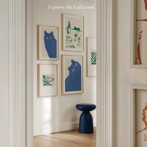 May include: A collection of framed art prints featuring blue cat illustrations and green line drawings. The prints are displayed on a white wall, with a blue stool in the foreground. The text "Explore the Collection" is at the top.