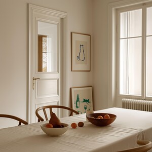 May include: A dining room scene with a large wooden table covered in a cream-colored tablecloth. A wooden bowl holds oranges and a pear. Two framed art prints of cats hang on the wall. A white door and window are also visible.
