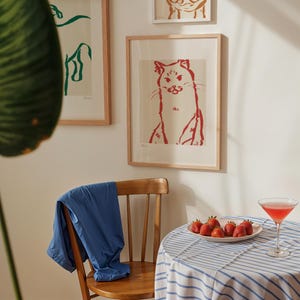 May include: A red cat art print in a wooden frame is displayed on a white wall, with other framed art. A blue jacket is draped over a wooden chair. A small table is set with strawberries and a cocktail.