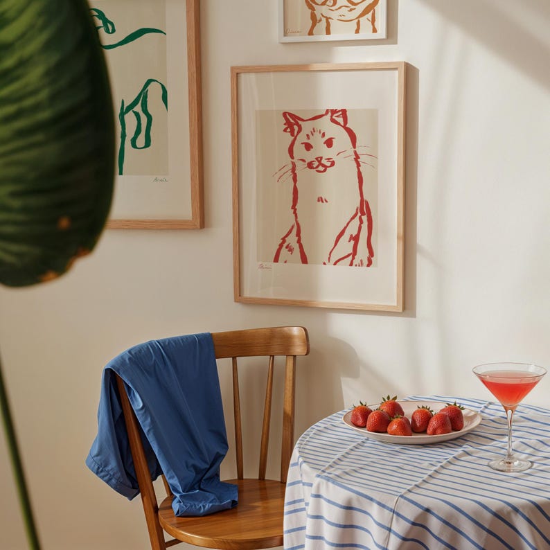 May include: A red cat art print in a wooden frame is displayed on a white wall, with other framed art. A blue jacket is draped over a wooden chair. A small table is set with strawberries and a cocktail.