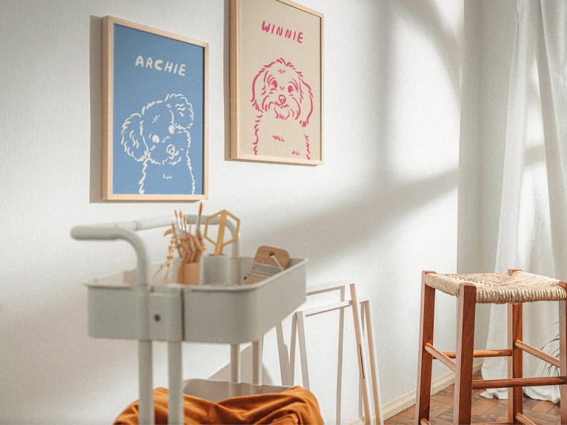 K&ouml;nnte beinhalten: Zwei gerahmte Hundeportr&auml;ts h&auml;ngen an einer wei&szlig;en Wand. Eines zeigt eine wei&szlig;e Illustration eines Hundes auf blauem Hintergrund mit dem Namen "Archie". Das andere hat eine rosa Hundeillustration auf beigem Hintergrund mit dem Namen "Winnie".