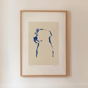 Puede incluir: Una impresión artística enmarcada con un dibujo lineal azul minimalista del perfil de un perro sobre un fondo crema. La obra de arte está encerrada en un marco de madera marrón claro, creando una estética simple y moderna. La impresión está montada en una pared blanca.