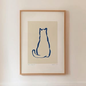 May include: Framed art print featuring a minimalist illustration of a cat. The cat is depicted in a simple, continuous blue line against a cream background. The artwork is encased in a light brown wooden frame, creating a clean and modern aesthetic.