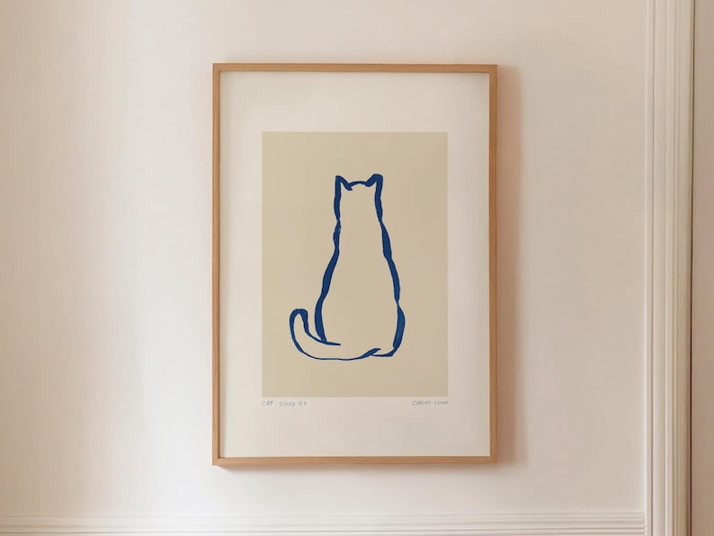 May include: Framed art print featuring a minimalist illustration of a cat. The cat is depicted in a simple, continuous blue line against a cream background. The artwork is encased in a light brown wooden frame, creating a clean and modern aesthetic.
