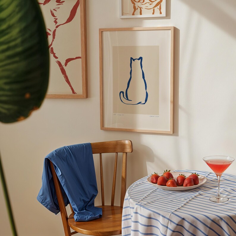 May include: A framed print of a blue cat silhouette hangs on a white wall. A wooden chair holds a blue jacket. A table with a blue and white striped tablecloth has a plate of strawberries and a cocktail.