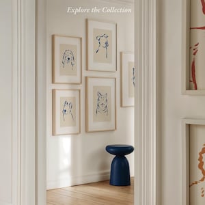 May include: A collection of framed dog and cat art prints with blue line drawings on a cream background. The prints are displayed on a white wall. A small, navy blue stool sits below the artwork. The text "Explore the Collection" is at the top.