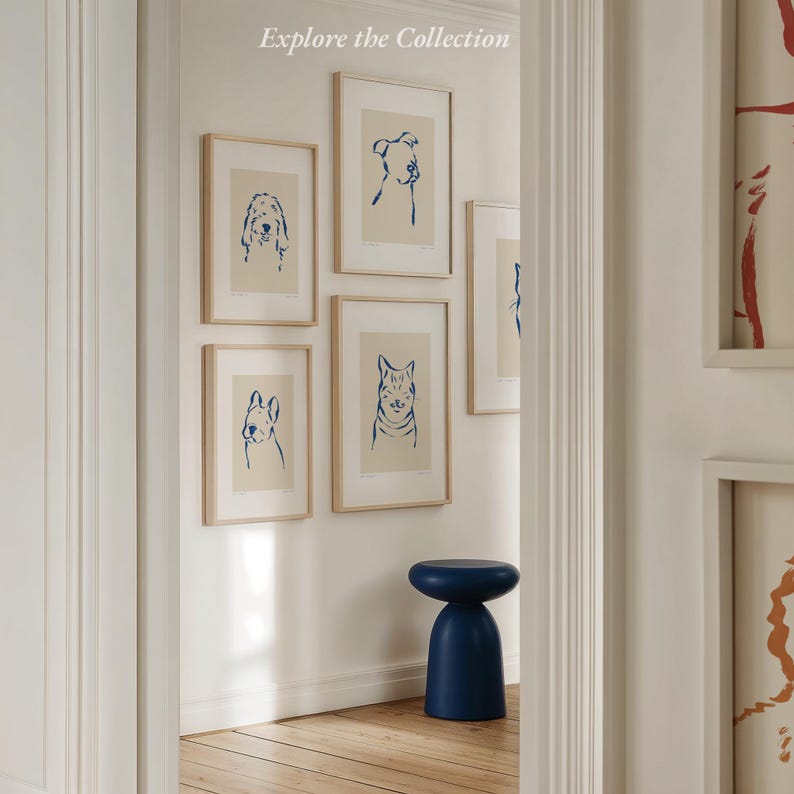 May include: A collection of framed dog and cat art prints with blue line drawings on a cream background. The prints are displayed on a white wall. A small, navy blue stool sits below the artwork. The text "Explore the Collection" is at the top.