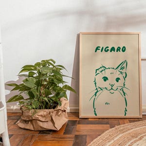 K&ouml;nnte beinhalten: Ein gerahmter Kunstdruck mit einer gr&uuml;nen Katzenillustration und dem Wort "FIGARO". Links befindet sich eine Topfpflanze in einer braunen Papiert&uuml;te. Das Kunstwerk steht auf einem Holzboden.