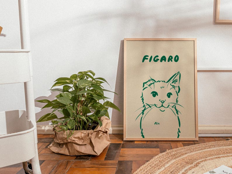 K&ouml;nnte beinhalten: Ein gerahmter Kunstdruck mit einer gr&uuml;nen Katzenillustration und dem Wort "FIGARO". Links befindet sich eine Topfpflanze in einer braunen Papiert&uuml;te. Das Kunstwerk steht auf einem Holzboden.