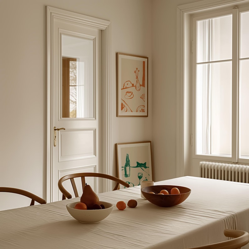 May include: A dining room scene featuring a wooden table set with a white tablecloth. A bowl of fruit and a wooden bowl are on the table. Two framed art prints hang on the wall. A white door and window are also visible.