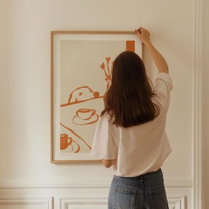 May include: A framed art print with a minimalist illustration in shades of orange and cream. The artwork depicts a dog, coffee cups, and a vase of flowers. The frame is a light wood color.