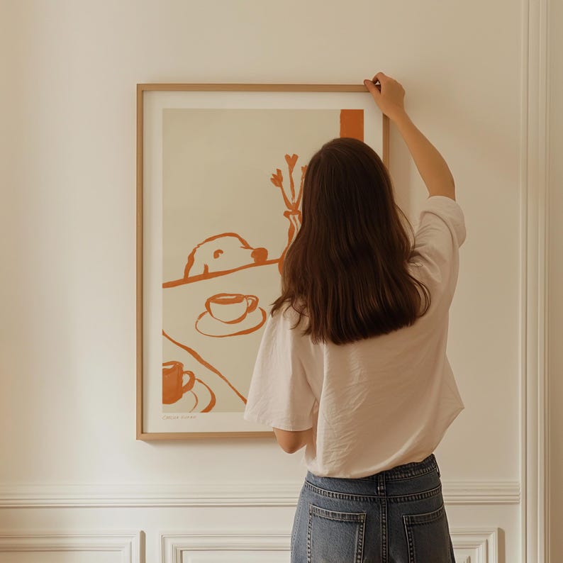 May include: A framed art print with a minimalist illustration in shades of orange and cream. The artwork depicts a dog, coffee cups, and a vase of flowers. The frame is a light wood color.
