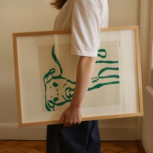 May include: A framed art print with a green abstract design on a cream background. The artwork is in a light-coloured wooden frame. The print is held by a person wearing a white t-shirt and blue jeans.