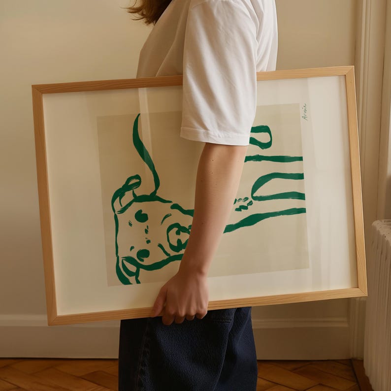 May include: A framed art print with a green abstract design on a cream background. The artwork is in a light-coloured wooden frame. The print is held by a person wearing a white t-shirt and blue jeans.