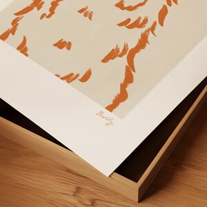 May include: Abstract art print with orange brushstrokes on a cream background, presented in a wooden frame. The artist's signature is visible. The print is angled, showing the frame's construction. The frame is made of wood.
