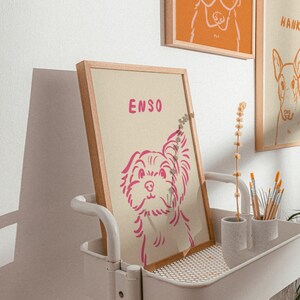 K&ouml;nnte beinhalten: Gerahmter Druck mit einer rosa Hundeillustration und dem Namen "ENSO". Das Kunstwerk wird auf einem wei&szlig;en Wagen mit anderen gerahmten Hundeportr&auml;ts im Hintergrund ausgestellt. Minimalistischer Stil, heller Holzrahmen.