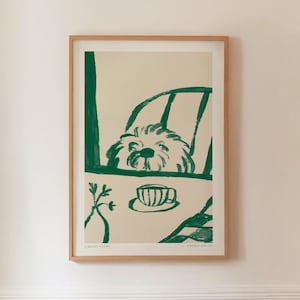 May include: A framed art print featuring a green and cream illustration. The artwork depicts a dog peeking over a table with a teacup and a plant. The print is in a wooden frame.