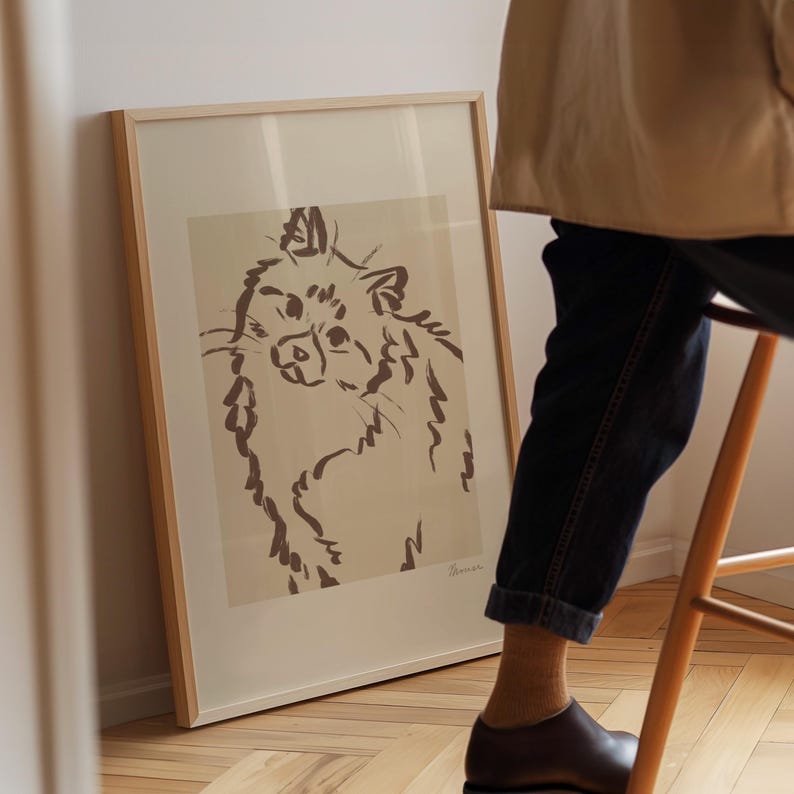 May include: A framed art print with a brown ink sketch of a cat. The artwork is in a light wood frame, leaning against a wall. The cat illustration has a whimsical, expressive style.