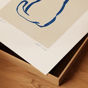 May include: Abstract art print featuring a blue line drawing on a beige background. The artwork is signed "Chelsea Kuran" in blue ink. The print is displayed in a wooden frame.