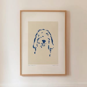 Puede incluir: Impresión enmarcada que presenta un dibujo lineal azul de la cara de un perro sobre un fondo crema. La obra de arte está en un marco de madera marrón claro. Los rasgos del perro están representados con líneas expresivas.