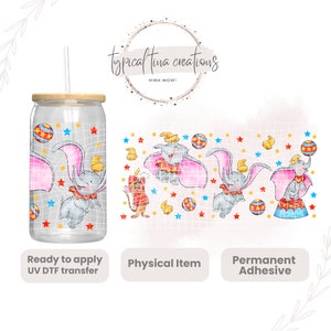 Puede incluir: Un vaso de vidrio transparente con una tapa de madera y una pajita. El vaso tiene un diseño de un elefante de dibujos animados con una capa rosa y una pelota roja y azul. El diseño se repite en una hoja separada de papel de transferencia transparente. El texto "typicaltina creations" y "MIRA WOW!" es visible en la imagen. El texto "Ready to apply UV DTF transfer", "Physical Item" y "Permanent Adhesive" también es visible en la imagen.