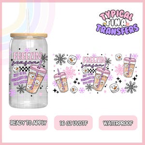 May include: A clear glass can-shaped cup with a wooden lid and a straw. The cup features a design with the text "FREEZIN season" and a cartoon image of a snowman in a drink. The design also includes snowflakes and the text "BUT STILL DRINKING AN ICED COFFEE".