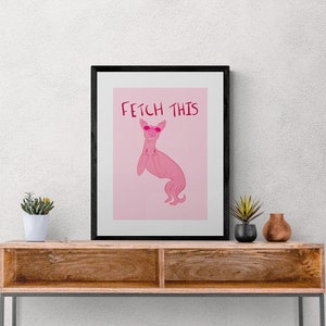 Könnte beinhalten: Ein schwarzer gerahmter Druck mit rosa Hintergrund, auf dem eine rosa Comic-Katze mit Sonnenbrille und dem Text "Fetch This" zu sehen ist. Der Druck wird auf einem Holz-Konsolentisch mit einem weißen Korb aus Seil auf der unteren Ablage präsentiert.