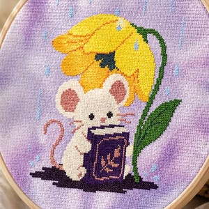May include: A circular wooden embroidery hoop containing a cross-stitch design on light purple fabric. The design features a white mouse holding a purple book while standing under a large yellow flower during a light blue rain shower.