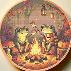 Peut inclure: Deux grenouilles vertes assises autour d'un feu de camp dans un décor forestier. Les grenouilles font griller des guimauves sur des bâtons. La scène est entourée de feuilles d'automne et de citrouilles.