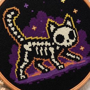 May include: A circular wooden embroidery hoop containing a cross-stitch design of a white cat skeleton on a black background. The cat is surrounded by purple clouds and small yellow and purple stars, with an orange outline around the cat's body.