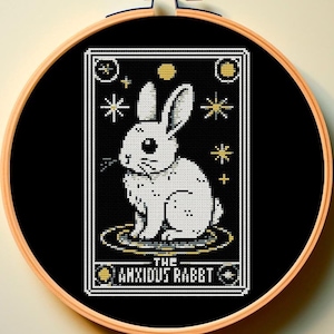 Puede incluir: Un patrón de punto de cruz en blanco y negro de un conejo blanco con un halo amarillo, rodeado de estrellas y lunas. El texto "The Anxious Rabbit" está debajo del conejo.