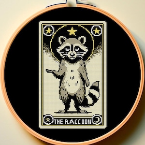 May include: A black and white cross-stitch pattern of a raccoon with a star above its head. The pattern is framed in a wooden hoop and the text "THE RACCOON" is below the raccoon.