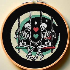 May include: Cross-stitch embroidery hoop with a black background featuring two skeletons on a swing with a red heart between them. The swing is suspended from a crescent moon with stars.