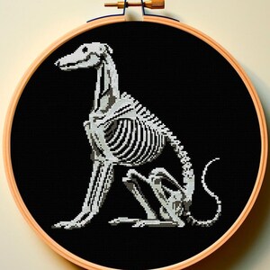 May include: A black and white cross-stitch embroidery of a dog skeleton sitting on a black background. The embroidery is framed in a wooden hoop.