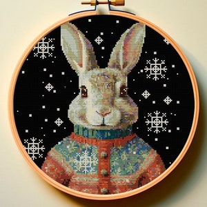 May include: A cross-stitch embroidery of a brown rabbit wearing a colorful sweater with snowflakes on a black background.