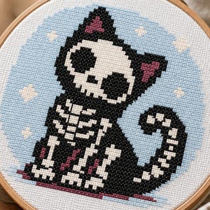 May include: A cross-stitch embroidery piece in a circular wooden hoop featuring a black cat with a white skeletal pattern on its body and head, set against a light blue circular background with small white stars, rendered in a pixelated style.